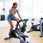 Jazzmyne Rain, a young Black woman with long red curly hair, confidently rides a stationary bike during her lifestyle branding session. She is wearing a sleek blue sports outfit, friendly expression, lifestyle, brand photoshoot.