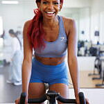 Jazzmyne Rain, a young Black woman with long red curly hair, confidently rides a stationary bike during her lifestyle branding session. She is wearing a sleek blue sports outfit, friendly expression, lifestyle, brand photoshoot.