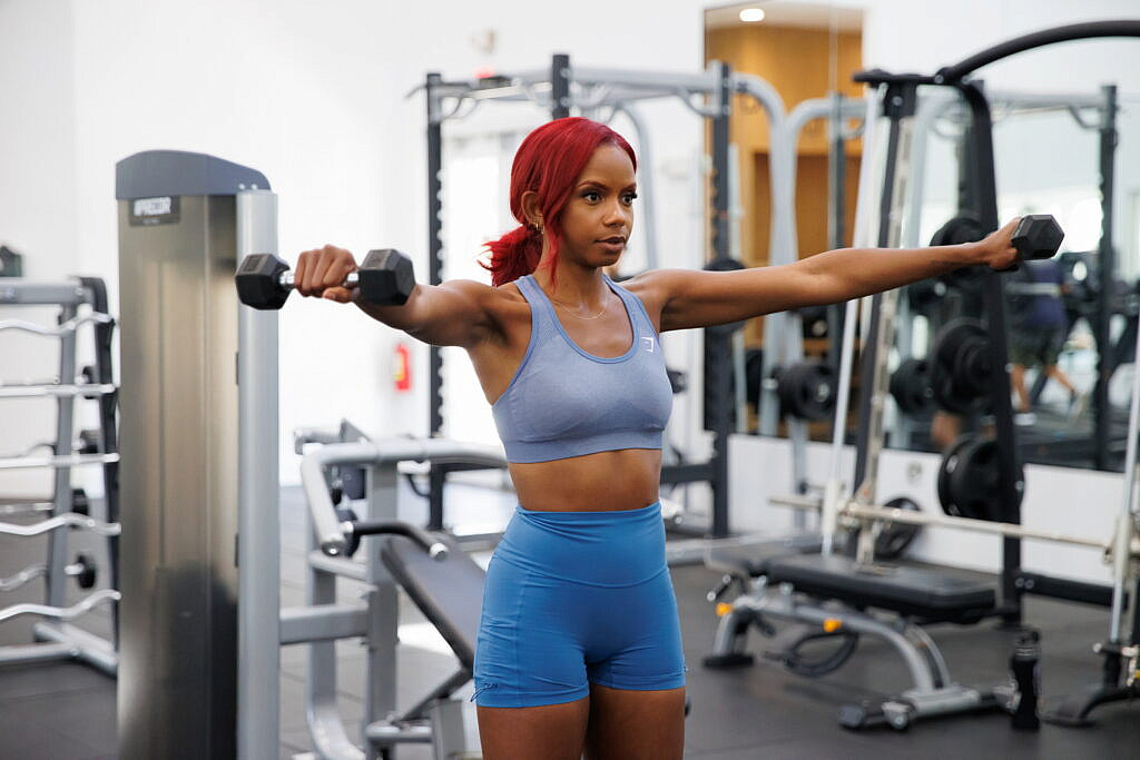 Jazzmyne Rain, a young Black woman with long red curly hair, confidently lifts dumbbells during her lifestyle branding session. She is wearing a sleek blue sports outfit, friendly expression, lifestyle, brand photoshoot.