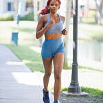 Jazzmyne Rain, a young Black woman with long red curly hair, confidently jogs along a path during her lifestyle branding session. She is wearing a sleek blue sports outfit, friendly expression, lifestyle, brand photoshoot.