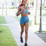 Jazzmyne Rain, a young Black woman with long red curly hair, confidently jogs along a path during her lifestyle branding session. She is wearing a sleek blue sports outfit, friendly expression, lifestyle, brand photoshoot.